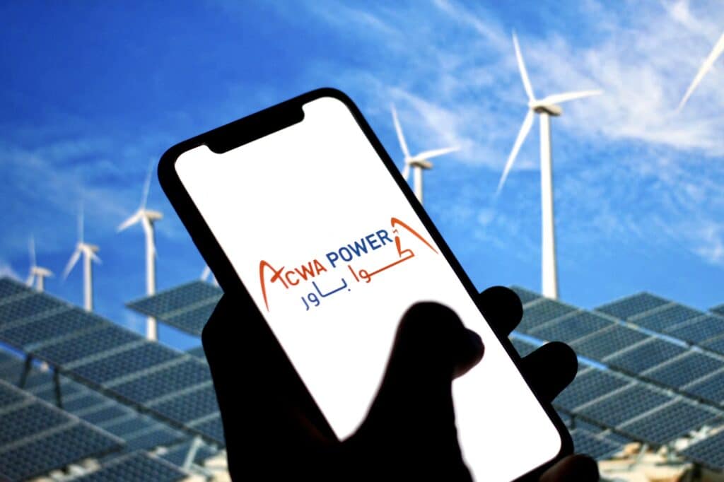 Логотип ACWA Power на экране смартфона на фоне солнечных батарей и ветряных турбин